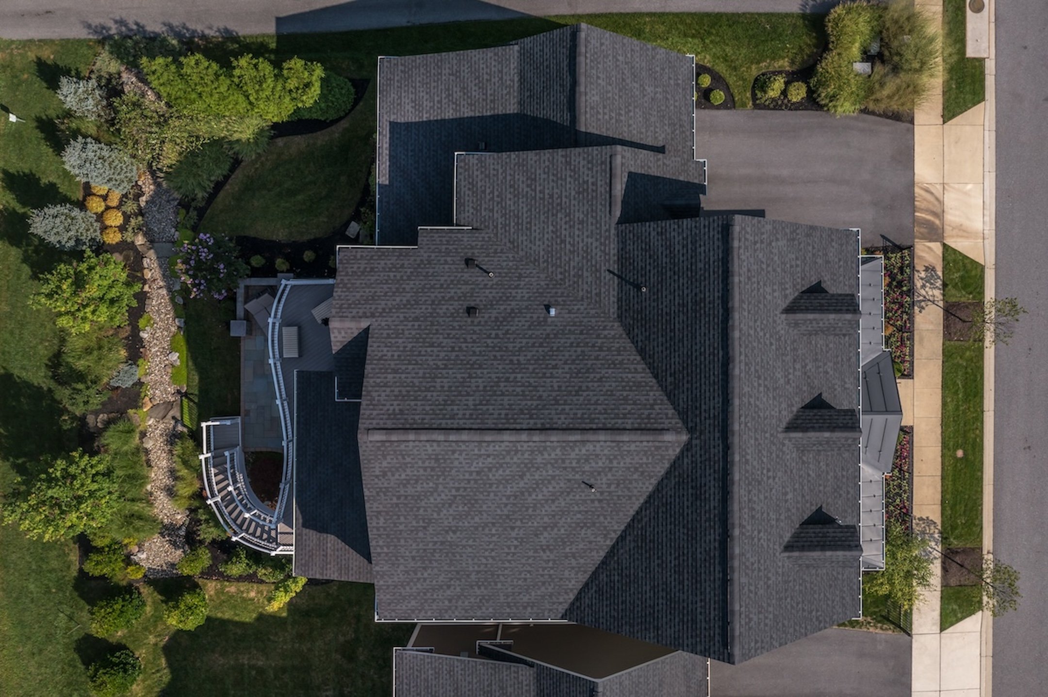Aerial view of a large house with dark gray roofing, surrounded by green lawns, trees, shrubs, and paved driveways on both sides. The property is bordered by roads and garden landscaping.