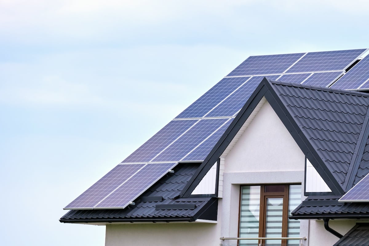A modern house with dark gray roof tiles has several large solar panels installed on its sloped roof, with a window and balcony visible below, against a light blue sky.