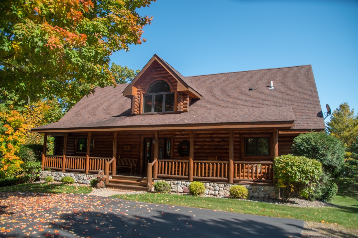 A log cabin with a stone foundation and a covered front porch sits under a clear blue sky, surrounded by green and autumn-colored trees, with fallen leaves scattered on the ground.