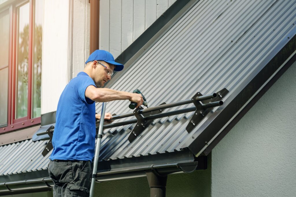 A worker in a blue shirt and cap uses a power drill to install a metal railing on a sloped metal roof of a house.