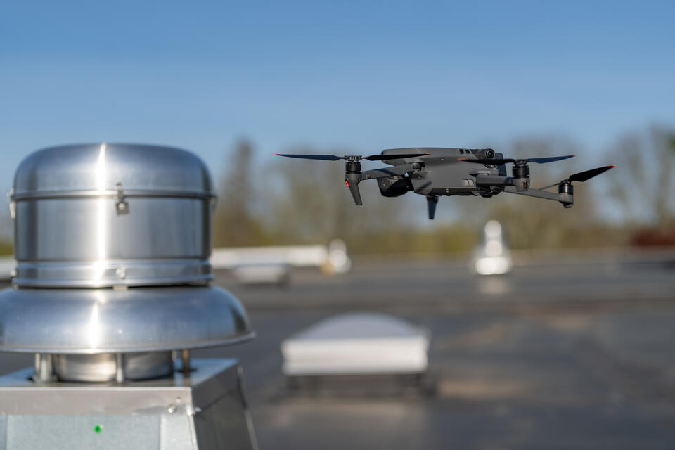 A black drone hovers above a flat rooftop near a silver ventilation unit, with a blurred background of trees and blue sky.