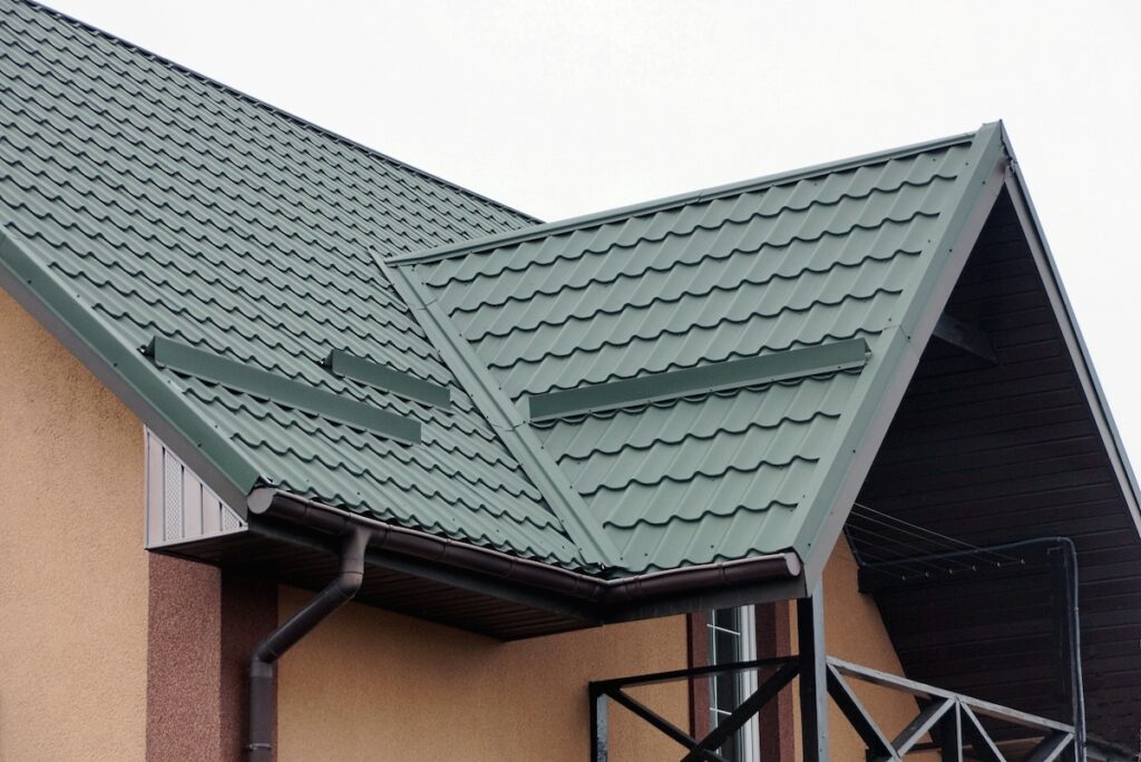 Green metal tile roof panels on a modern house with tan and brown walls, multiple roof slopes, rain gutters, and a covered entrance visible.