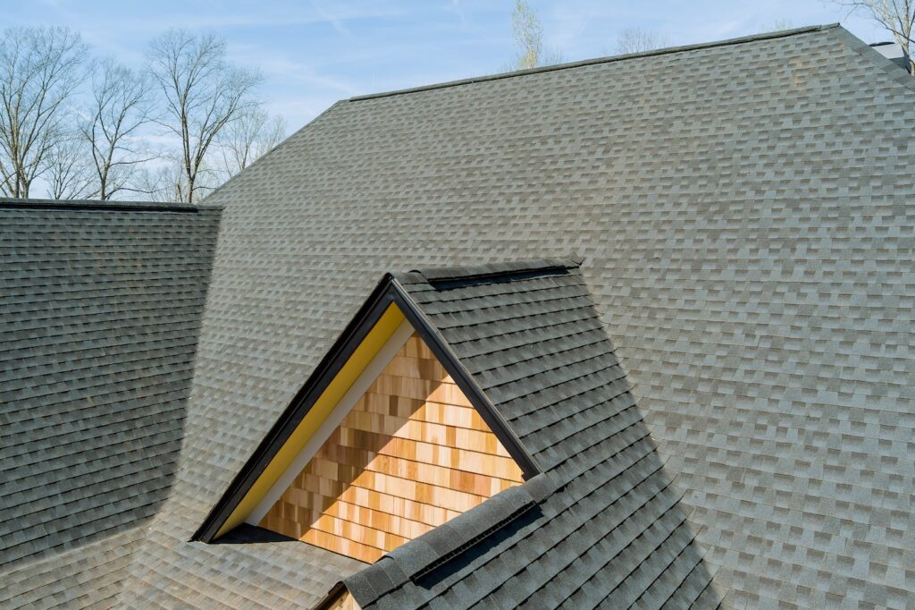 A close-up view of a roof with dark gray asphalt shingles, featuring a dormer with yellow trim and wood siding. Leafless trees are visible in the background under a clear sky.