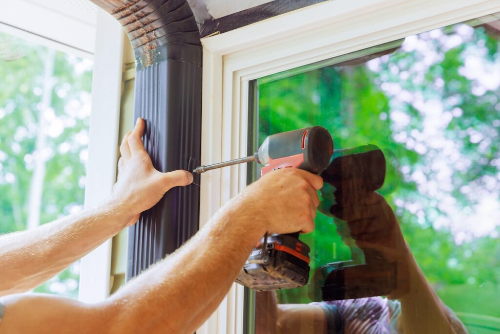 A person uses a power drill to attach a black drainpipe to the exterior wall next to a window, with reflections of trees visible in the glass.