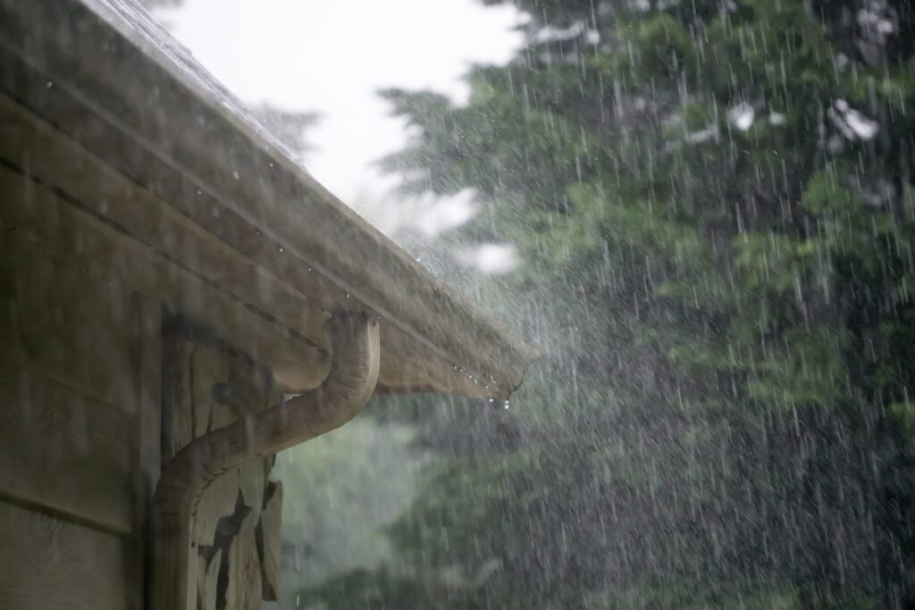 Rain pours heavily from the roof and gutters of a house, with water streaming down the side. Trees in the background appear blurred from the downpour, creating a misty, overcast atmosphere.