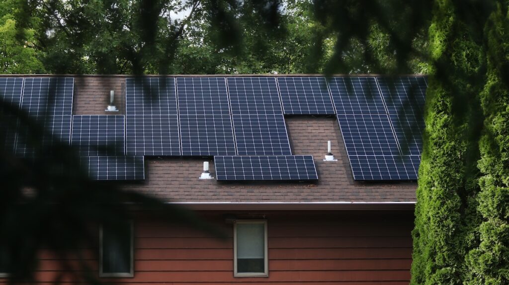 Solar panels are installed on the roof of a house, surrounded by green trees. The roof has vents that the solar panels are arranged around, and the house exterior is a reddish-brown color.