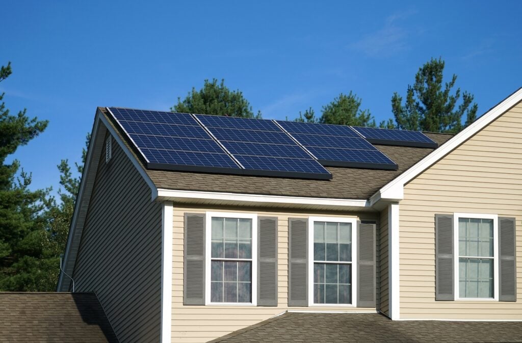A beige house with gray shutters has solar panels installed on its roof. The sky is clear and blue, and tall green trees are visible in the background.