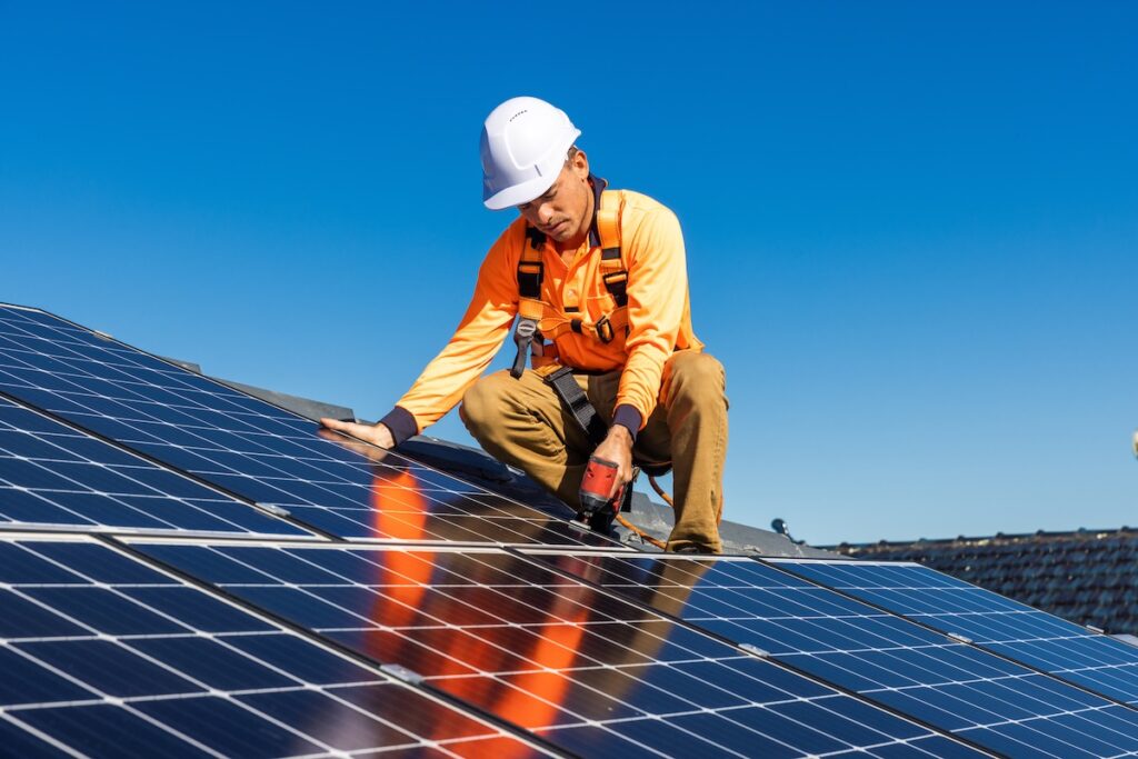 A worker in a hard hat and safety harness installs solar panels on a rooftop under a clear blue sky, using a cordless drill.