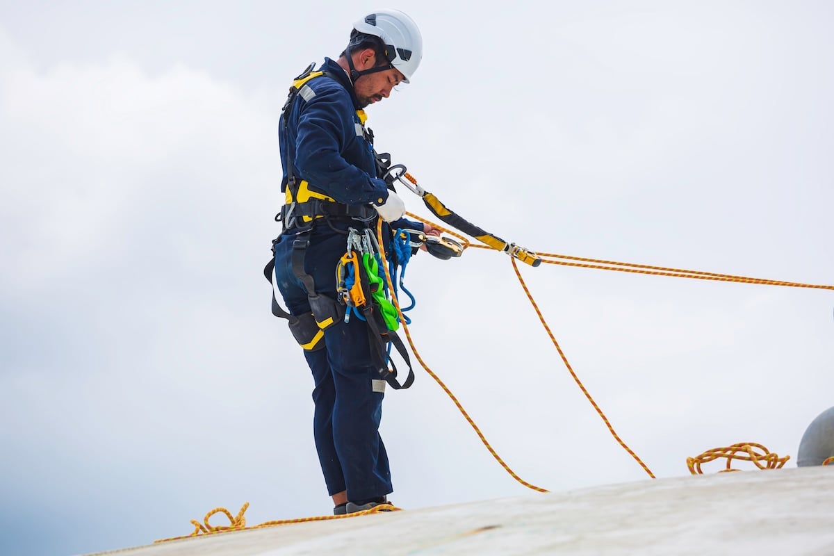A worker wearing safety gear and a helmet is using ropes and harnesses while standing on a sloped surface, performing maintenance or inspection work at height against a cloudy sky.