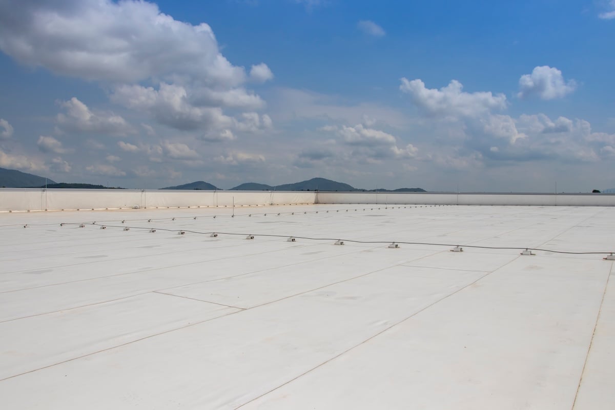 A large, flat white commercial roof with clear seam lines sits under a bright blue sky with scattered clouds—showcasing the results of a recent commercial flat roof replacement. Distant hills are visible along the horizon.