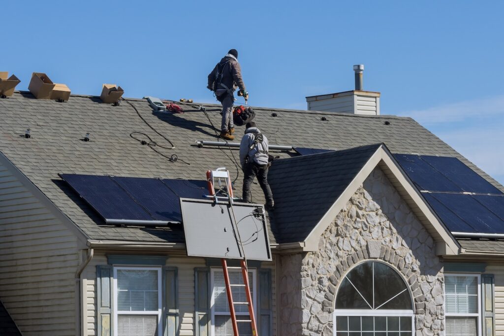 Two workers wearing safety gear install solar panels on the roof of a beige house with a stone facade. Tools and boxes are scattered on the roof, and a red ladder leans against the house.