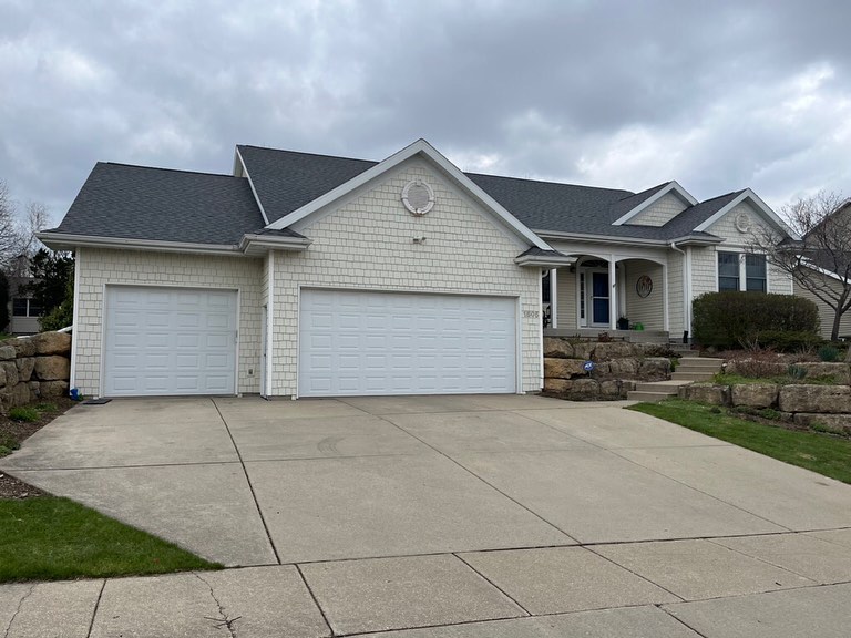 A large, light-colored suburban house in Chenequa features three white garage doors, a wide driveway, and landscaping with stone walls and shrubs. Its roofing showcases a dark gray finish above a front porch with columns under an overcast sky.