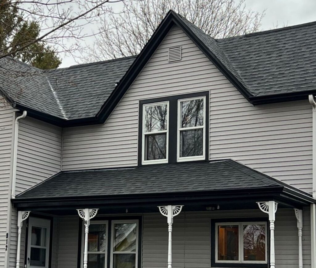 A two-story house with gray siding, black trim, white framed windows, and a covered front porch supported by decorative white columns. Quality roofing Brookfield work complements the style. Leafless trees stand in the background under a cloudy sky.