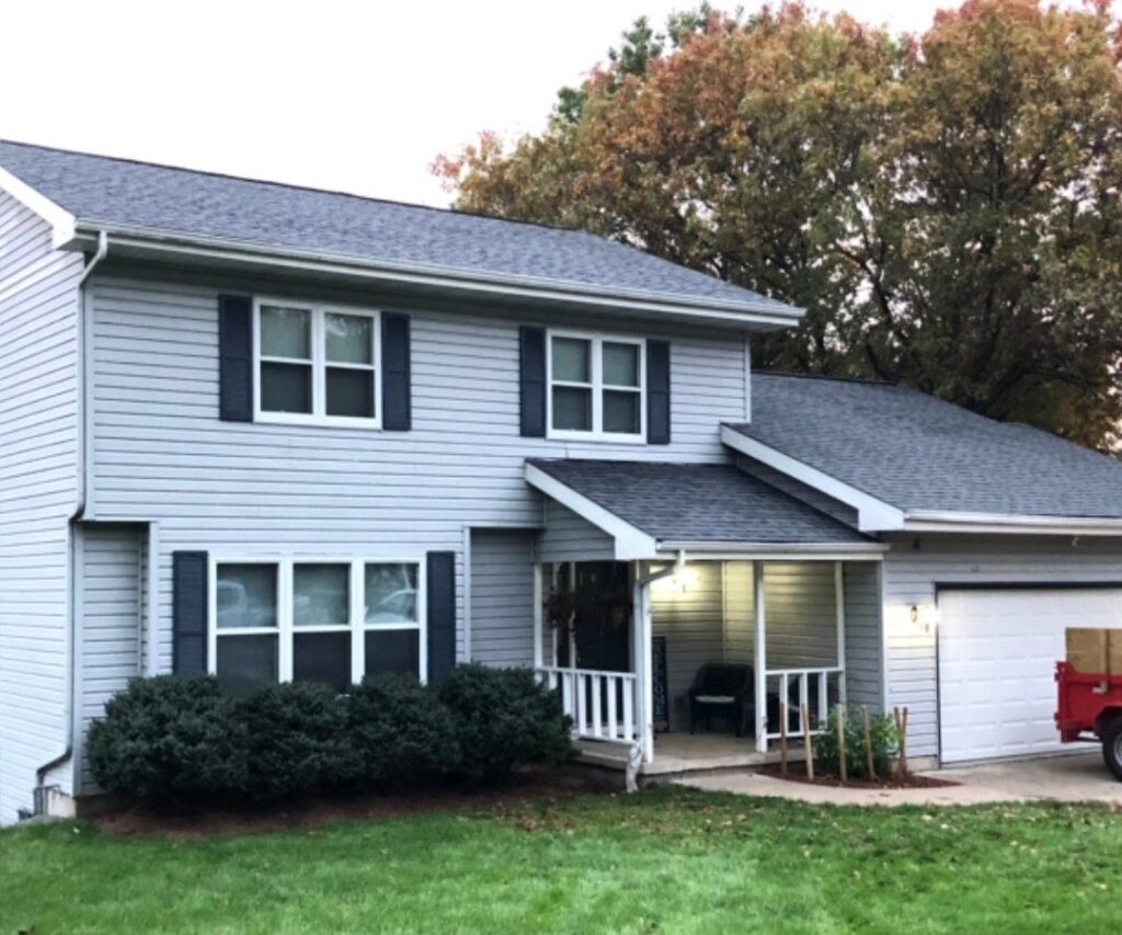 Two-story gray house with black shutters, a covered front porch, and an attached garage. Bushes line the front, a red dumpster sits in the driveway—perfect for recent roofing Delafield projects. Autumn trees add color to the background.