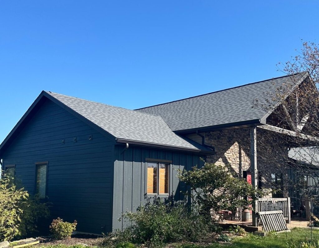A modern house with dark gray siding and a sloped roof, expertly crafted by roofing Pewaukee professionals, stands under a clear blue sky. Bushes and greenery add charm to the yard beside the house.