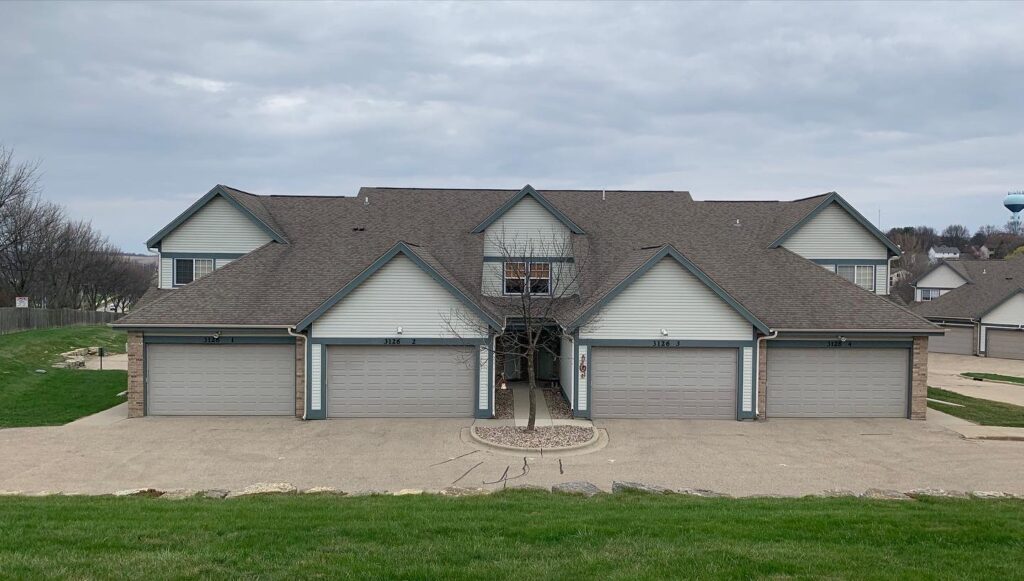 A duplex-style residential building with gray siding and brown roofs, featuring four garages and a small tree in front. Set under a cloudy sky with green grass in the foreground, it displays quality roofing Waukesha residents appreciate.