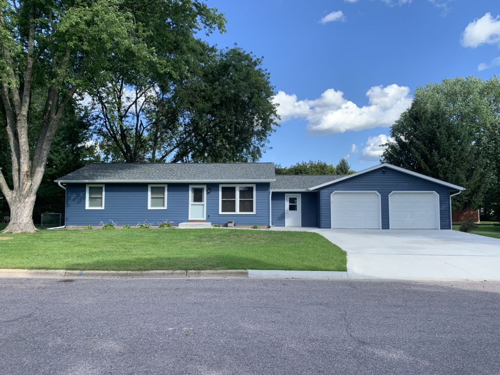 A single-story blue house with white trim features a new roofing Oconomowoc style, a large lawn, and a driveway to a double garage. Trees surround the property under a partly cloudy sky.