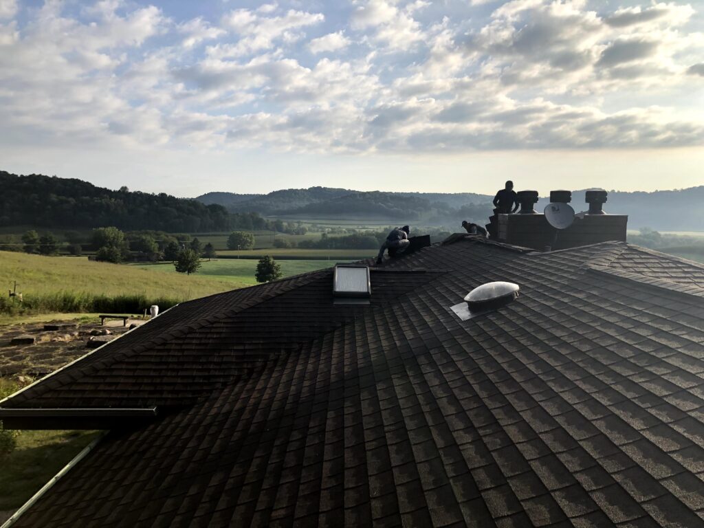 Two people work on a large roof under a partly cloudy sky, with fields, trees, and rolling hills visible in the background on a sunny day.