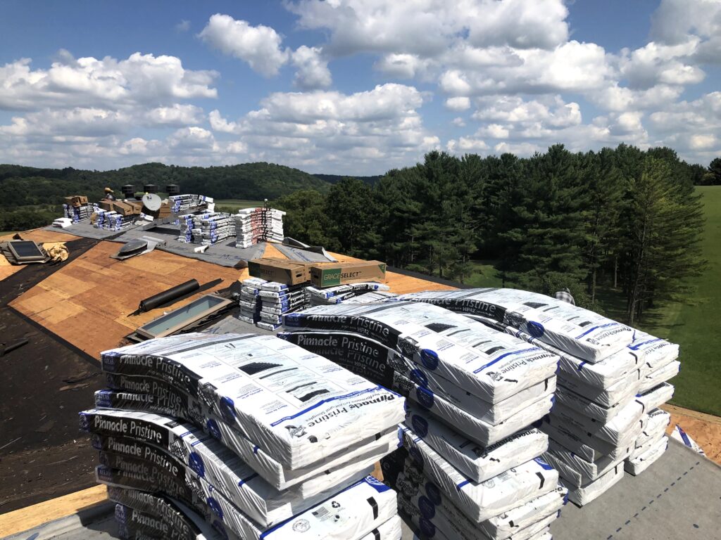 Stacks of roofing materials, including Pinnacle Pristine shingles, are piled on a roof under construction with open wood areas. In the background, there are trees, green fields, and a partly cloudy sky.