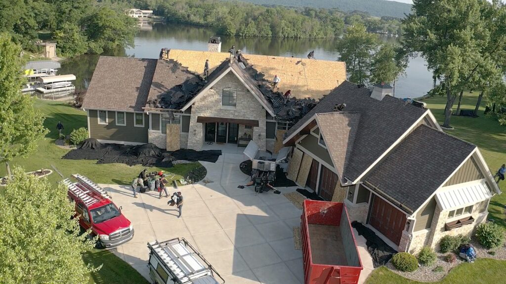 Aerial view of a large lakeside house undergoing roof repair. Workers are on the roof, some shingles are removed, and roofing materials are scattered. Construction vehicles and equipment are in the driveway. Trees and water in the background.