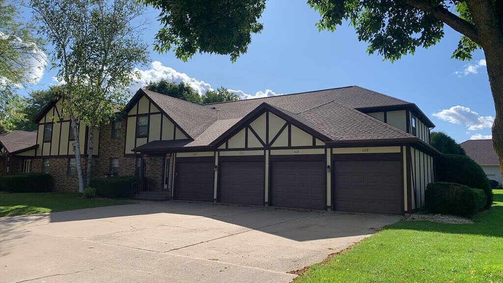 A large suburban house with Tudor-style architecture, three brown garage doors, brick and cream-colored exterior, quality roofing Milwaukee style, a spacious driveway, green lawn, and trees under a sunny blue sky with scattered clouds.