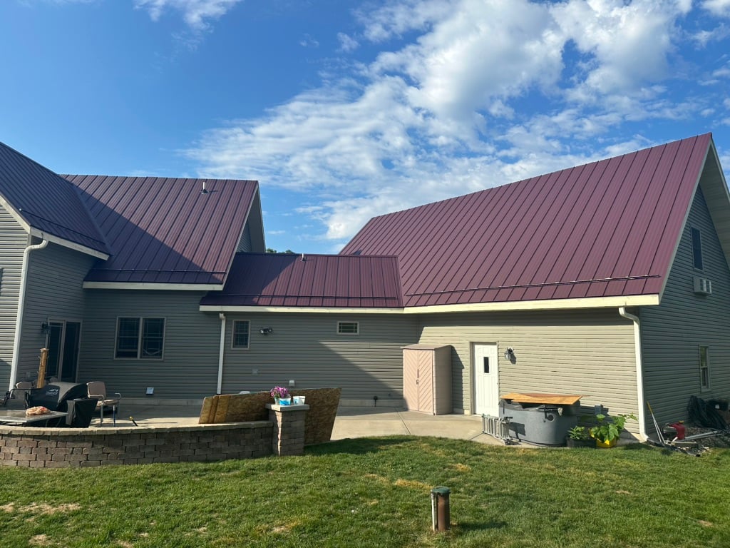 A large beige house with a red metal roof stands under a blue sky with scattered clouds. There is a patio area with outdoor furniture, a small shed, and various yard items on green grass.