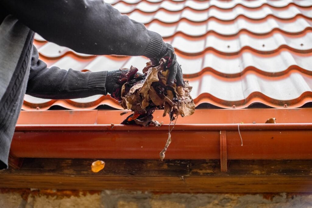 A person wearing black gloves clears dry leaves from a red gutter attached to a house with a red tile roof.