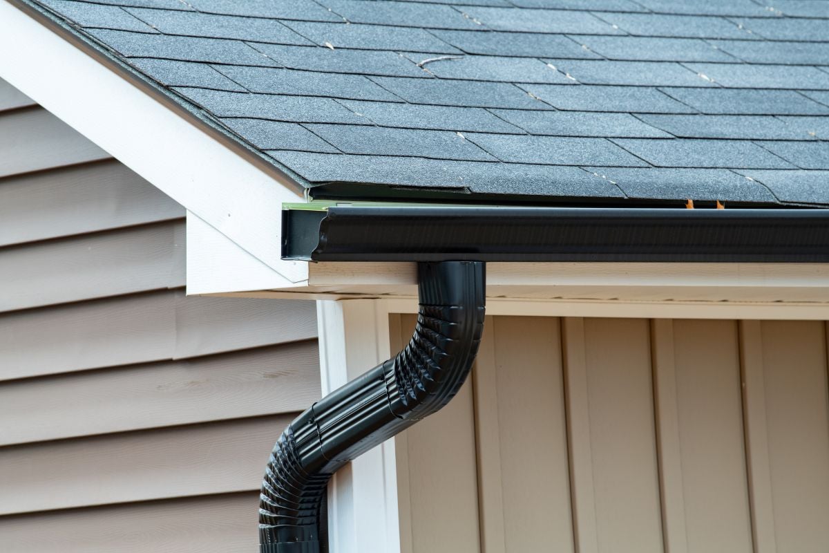 Close-up of the corner of a house roof with black shingles, beige siding, and a black rain gutter system with a downspout attached.