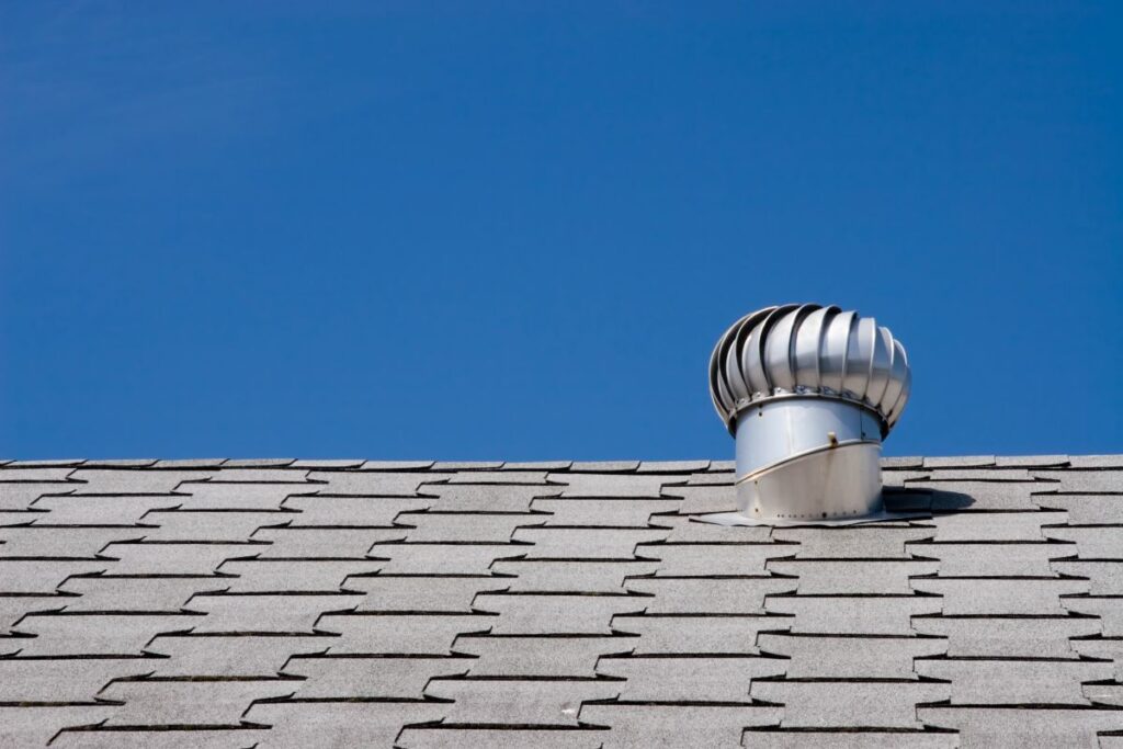 A silver metal roof vent sits on top of an asphalt shingle roof under a clear blue sky.