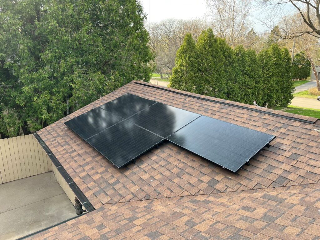 Three black solar panels installed on the brown shingled roof of a house, surrounded by green trees and a paved driveway below.
