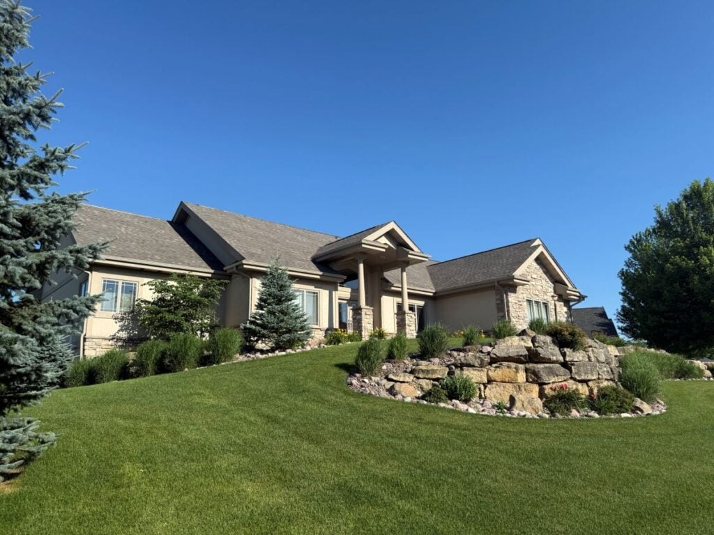 A beige house with a gabled roof by roofing Brookfield sits on a green, sloping lawn. Stone accents, landscaping with bushes, rocks, and evergreen trees complete the scene under a clear blue sky.