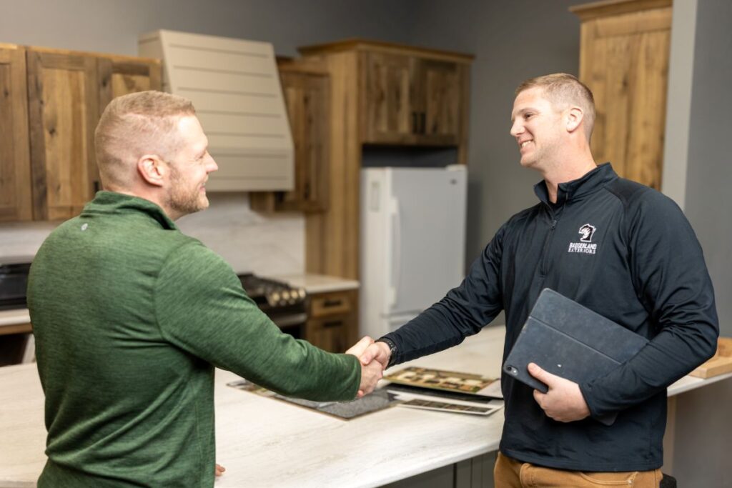 Two men are shaking hands in a modern kitchen. One man holds a folder, and they both appear to be smiling, suggesting a positive interaction or business agreement. Wooden cabinets and a white refrigerator are in the background.