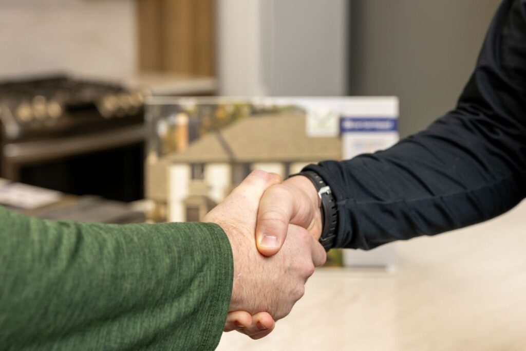 Two people shaking hands indoors, with blurred background showing what appears to be a kitchen and a house brochure, suggesting a real estate or business agreement.
