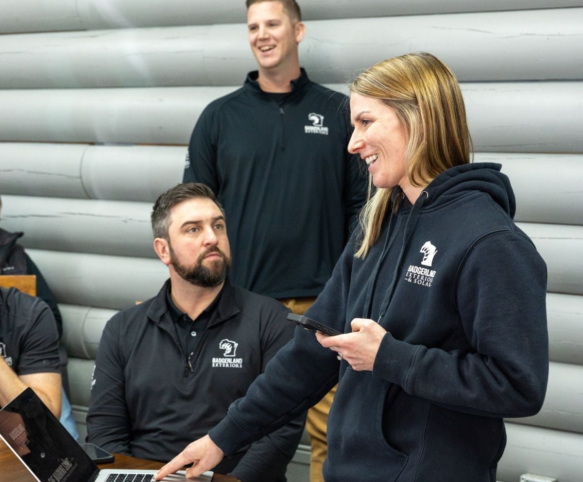 A woman in a hoodie smiles while holding a phone and pointing at a laptop, standing with three colleagues in front of a gray wooden wall. Two men sit at a table, and one man stands smiling in the background.