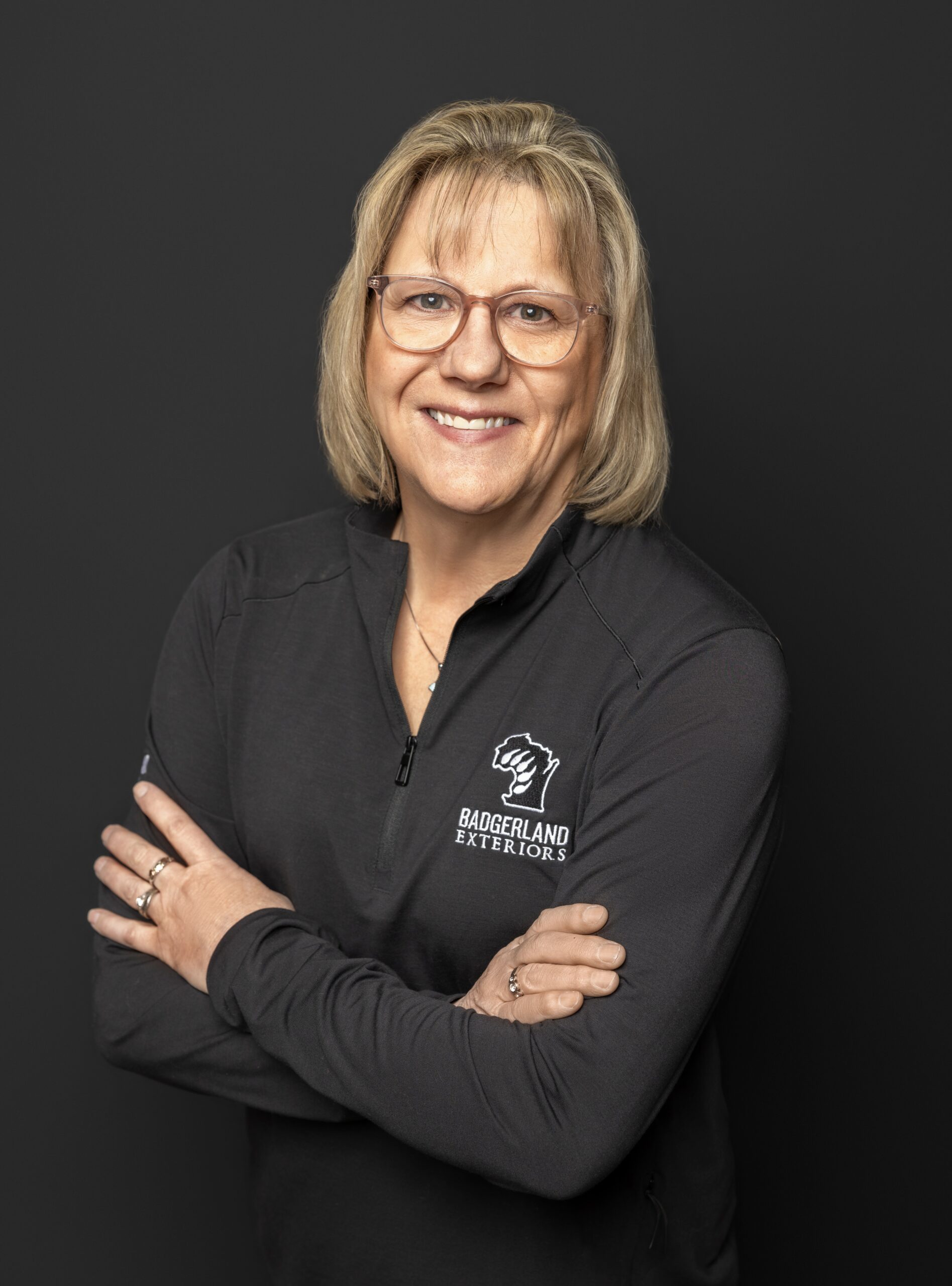 A smiling woman with blonde hair and glasses stands with her arms crossed, wearing a black zip-up shirt with the Baggerland Interiors logo on the chest, in front of a plain dark background.