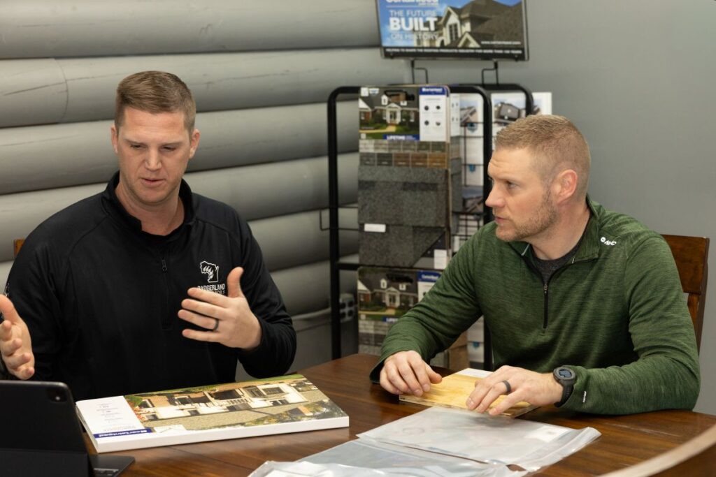 Two men sit at a table discussing home plans and material samples. One man gestures while speaking, and the other listens, holding a sample board. Brochures and sample boards are on the table, with displays in the background.