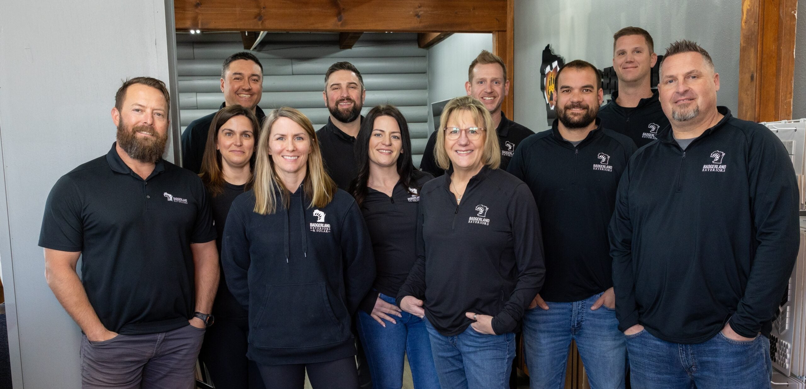 A group of eleven people, five women and six men, stand indoors smiling at the camera. They are wearing matching black shirts with a logo, suggesting they are part of the same team or organization.