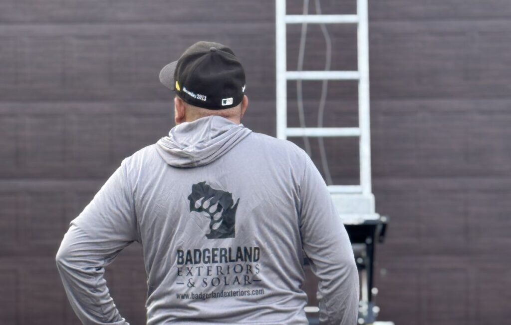 A person wearing a gray hoodie and black cap stands with their back to the camera. The hoodie has a logo with a fist and text reading "Badgerland Exteriors & Solar" and a website. A metal ladder is in the background.
