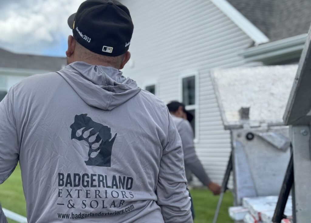 A person wearing a gray "Badgerland Exteriors & Solar" hoodie stands outside near a house, with another worker and equipment visible in the background. The focus is on the hoodie logo.