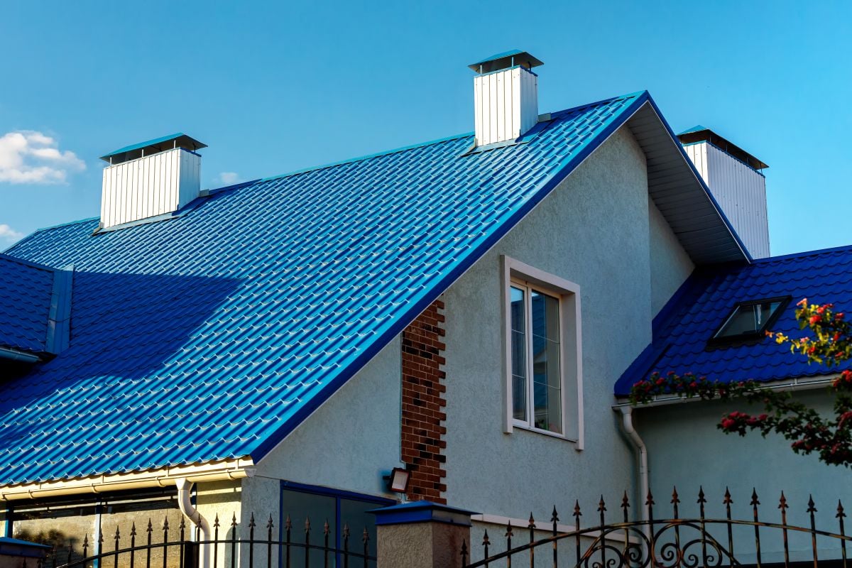 benefits of metal roofing house with a blue metal roof, white-framed windows, and two white chimneys, set against a clear blue sky. A black fence and some greenery are visible in the foreground.