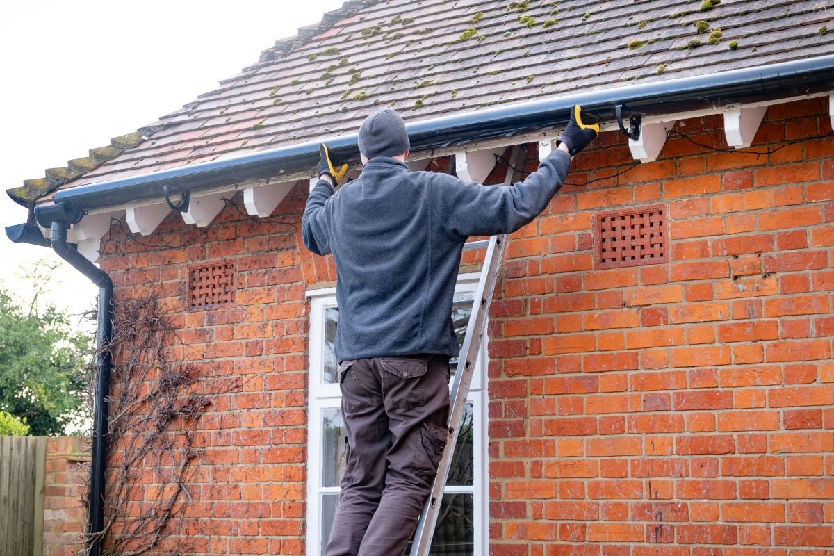 gutter replacement person standing on a ladder inspects or cleans the gutters of a brick house with a tiled roof, wearing gloves, a beanie, and a jacket, on a clear day.