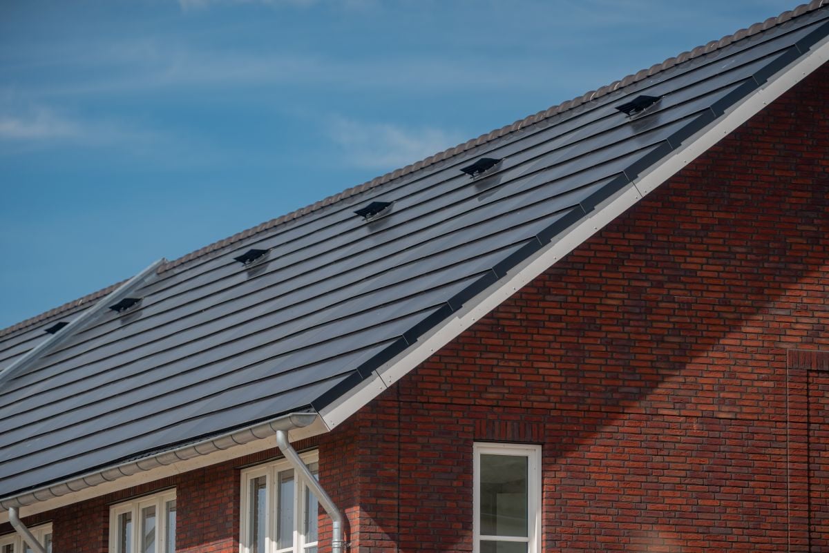gaf solar roof shingles Close-up of a sloped house roof with dark gray solar shingles, set against a blue sky. The brick exterior wall and white-framed windows are partially visible beneath the roof.
