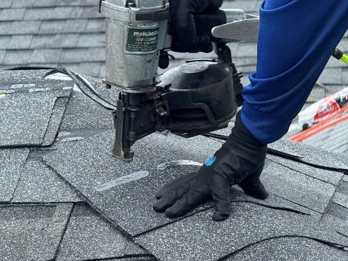roof replacement timeline person wearing black gloves uses a nail gun to install gray asphalt shingles on a roof. The shingles are layered, and roofing tools are visible, with part of the worker’s blue sleeve showing.