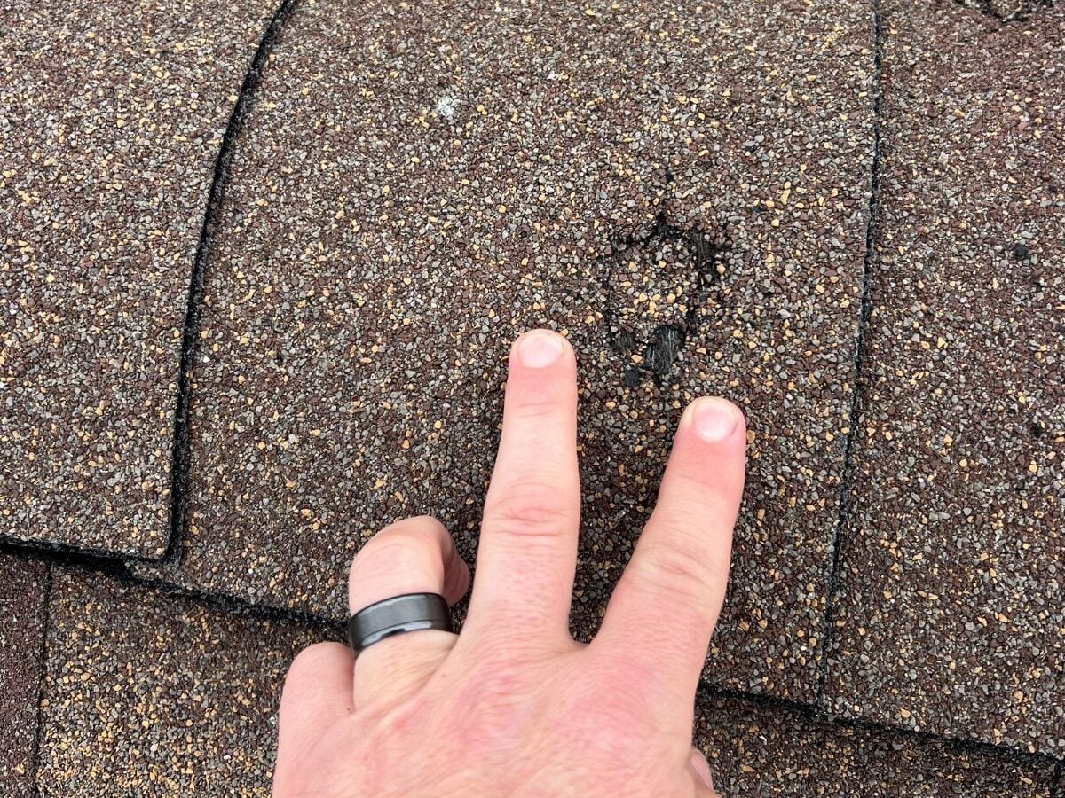 storm damage roof repair A hand with two fingers pointing at a small damaged spot on an asphalt shingle roof, highlighting an area where the shingles appear worn or dented.