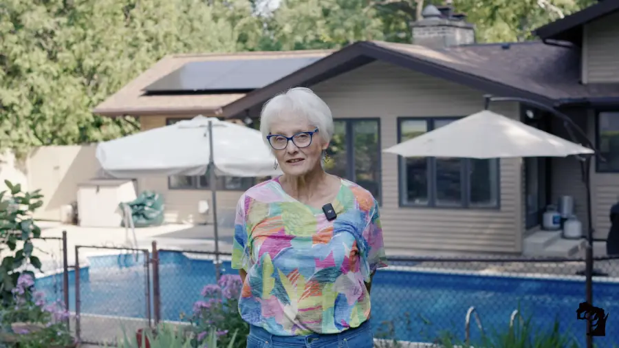 An older woman with short white hair and glasses stands in front of a backyard pool and house, wearing a colorful shirt and jeans, with greenery and umbrellas in the background.