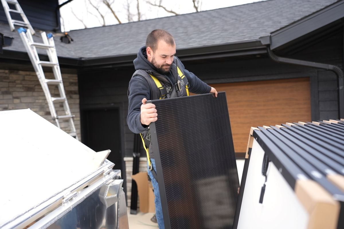 wisconsin solar incentives man wearing a safety harness and hoodie lifts a solar panel outside a house with dark siding and a brown garage door. A ladder leans against the roof and several solar panels are stacked nearby.