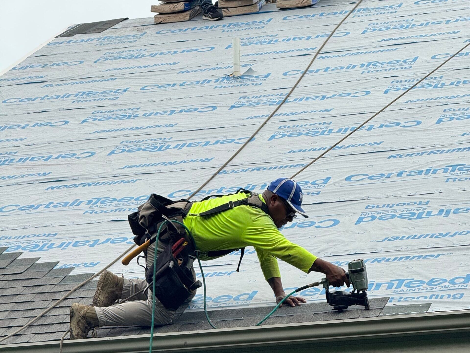 roofing companies middleton wi A construction worker wearing a safety harness and a neon shirt uses a nail gun to install shingles on a house roof. Roofing materials and tools are visible around him.
