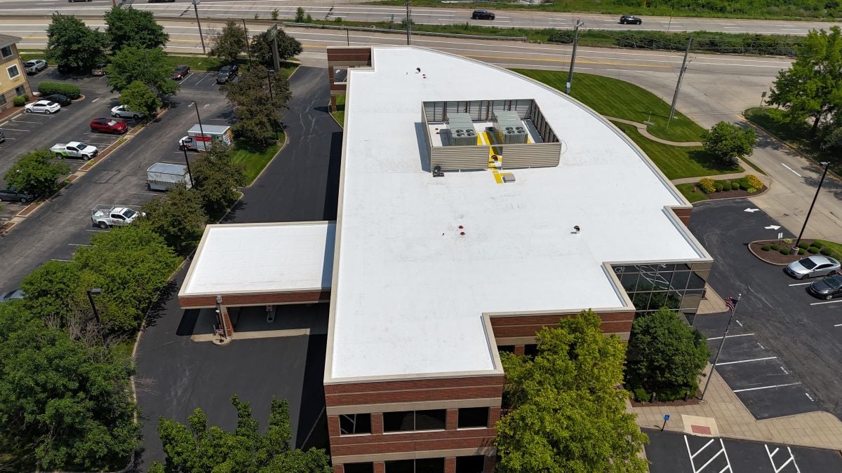 commercial roof replacement Aerial view of a modern commercial building with a white flat roof and HVAC units on top, surrounded by trees, parking lots, and nearby roads.
