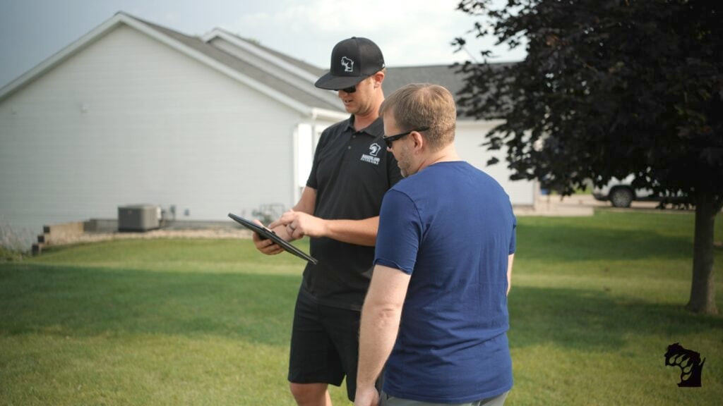 what is the most expensive part of replacing a roof in middleton wi Two men stand on a lawn near a house; one, wearing a black cap and shirt, points at a tablet while the other, in a blue shirt, looks on. A tree and part of a building are visible in the background.