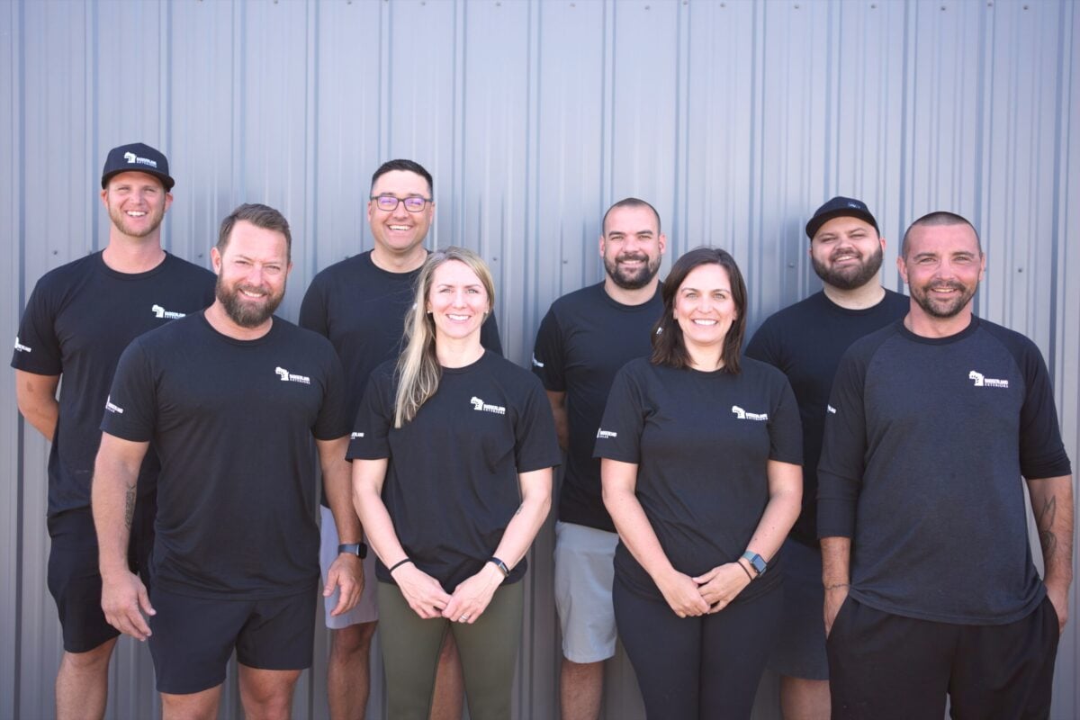 roofing companies verona wi Eight people stand in two rows, smiling in front of a corrugated metal wall. They all wear matching black shirts with a small white logo on the chest. The group appears friendly and casual.